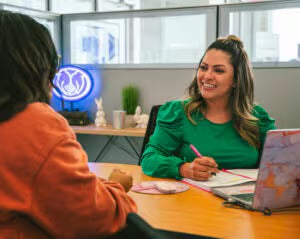 A customer service representative smiling and taking notes while speaking with a client across a desk.