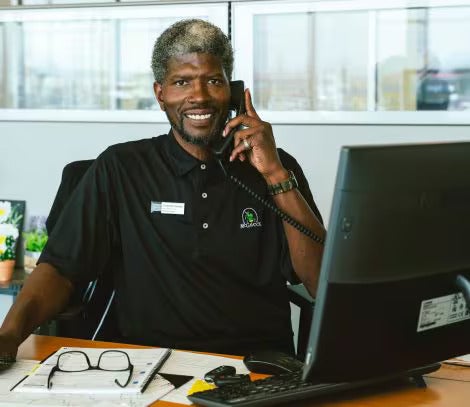 A smiling man sits at a desk with glasses, a keyboard, and paperwork, speaking on a phone.